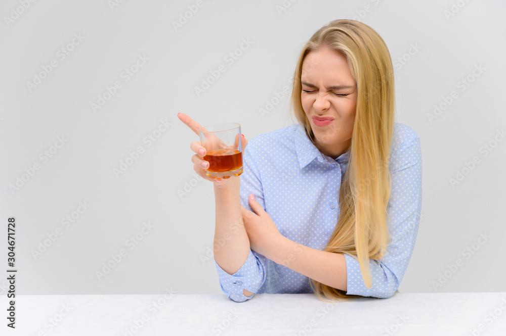 Concept woman with alcohol problems sits at a table with whiskey in a glass. Portrait of a beautiful blonde girl with excellent makeup with long smooth hair on a white background in a blue shirt. Concept woman with alcohol problems sits at a table with whiskey in a glass. Portrait of a beautiful blonde girl with excellent makeup with long smooth hair on a white background in a blue shirt.