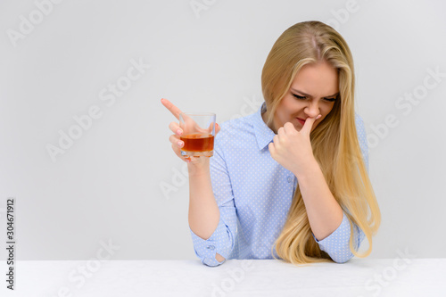 Concept woman with alcohol problems sits at a table with whiskey in a glass. Portrait of a beautiful blonde girl with excellent makeup with long smooth hair on a white background in a blue shirt.