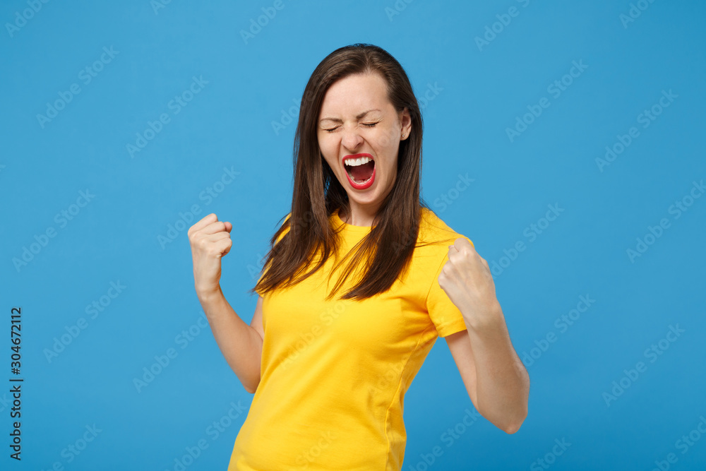 Fototapeta premium Happy young brunette woman girl in yellow t-shirt posing isolated on bright blue background. People lifestyle concept. Mock up copy space. Clenching fists like winner, keeping eyes closed, screaming.