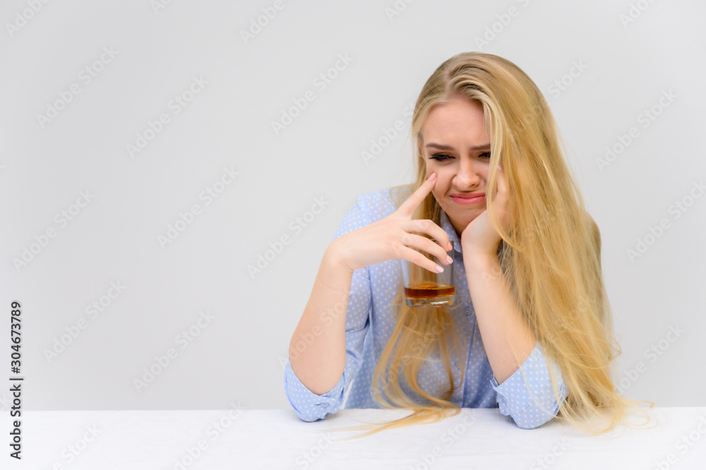 Concept woman with alcohol problems sits at a table with whiskey in a glass. Portrait of a beautiful blonde girl with excellent makeup with long smooth hair on a white background in a blue shirt. Concept woman with alcohol problems sits at a table with whiskey in a glass. Portrait of a beautiful blonde girl with excellent makeup with long smooth hair on a white background in a blue shirt.