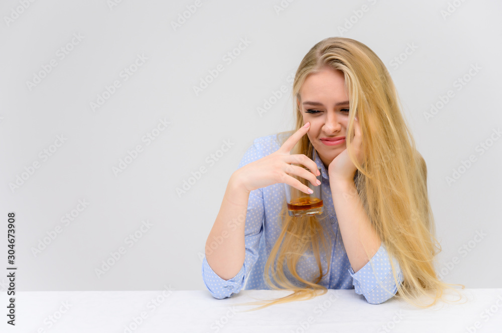 Concept woman with alcohol problems sits at a table with whiskey in a glass. Portrait of a beautiful blonde girl with excellent makeup with long smooth hair on a white background in a blue shirt. Concept woman with alcohol problems sits at a table with whiskey in a glass. Portrait of a beautiful blonde girl with excellent makeup with long smooth hair on a white background in a blue shirt.
