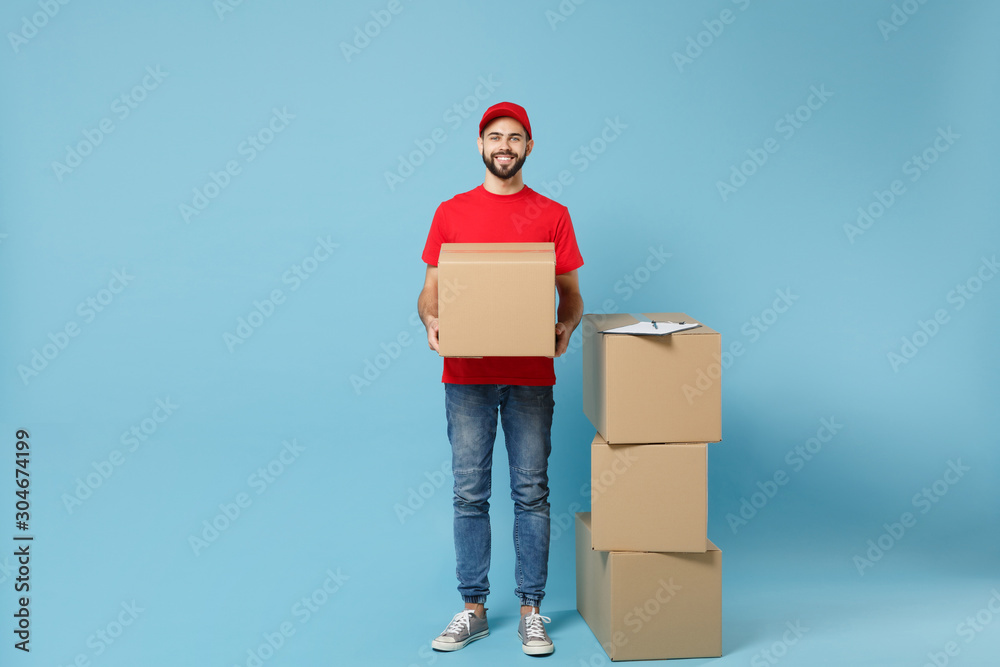 Delivery man in red uniform isolated on blue background, studio portrait. Male employee in cap t-shirt print working as courier dealer hold empty cardboard box. Service concept. Mock up copy space. Delivery man in red uniform isolated on blue background, studio portrait. Male employee in cap t-shirt print working as courier dealer hold empty cardboard box. Service concept. Mock up copy space.