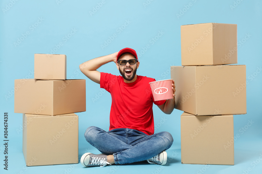 Delivery man in red uniform isolated on blue background, studio portrait. Male employee in cap t-shirt print working as courier dealer sit at empty cardboard box. Service concept. Mock up copy space. Delivery man in red uniform isolated on blue background, studio portrait. Male employee in cap t-shirt print working as courier dealer sit at empty cardboard box. Service concept. Mock up copy space.