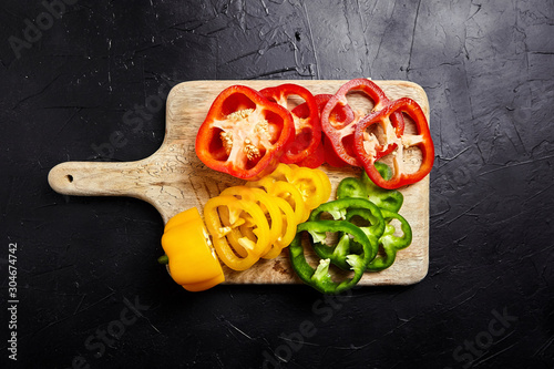 Cutting board with slices of red, green, yellow bell peppers on black background. Sliced sweet peppers in different colors on stone table, vegetable salad ingredient, cooking healthy food, top view photo