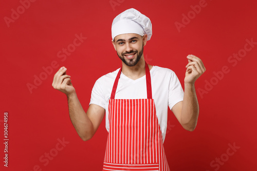 Bearded male chef cook or baker man in striped apron toque chefs hat posing isolated on red background. Cooking food concept. Mock up copy space. Rubbing fingers showing cash gesture asking for money.