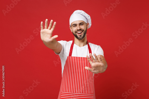 Cheerful bearded male chef cook or baker man in striped apron toque chefs hat posing isolated on red background. Cooking food concept. Mock up copy space. Stand with outstretched hands, showing palms.
