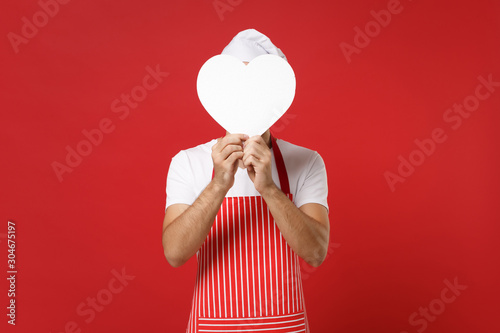 Young male chef cook or baker man in striped apron t-shirt toque chefs hat isolated on red background in studio. Cooking food concept. Mock up copy space. Covering face with white empty blank heart.