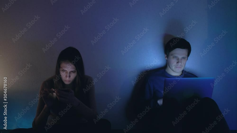 Bored young couple using smartphone and laptop at night. Concept of exhausted family relationships. Bored young couple using smartphone and laptop at night. Concept of exhausted family relationships.