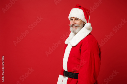 Side view of smiling elderly gray-haired mustache bearded Santa man in Christmas hat posing isolated on red background. New Year 2020 celebration holiday concept. Mock up copy space. Looking camera.