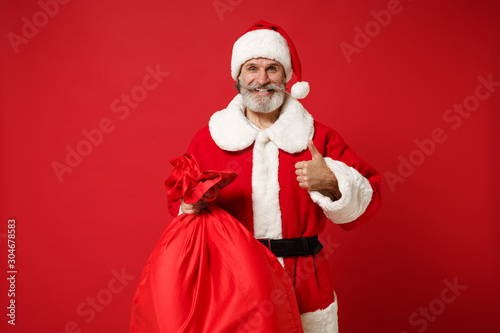 Elderly gray-haired mustache bearded Santa man in Christmas hat posing isolated on red background. New Year 2020 celebration holiday concept. Mock up copy space. Hold bag with gifts, showing thumb up.