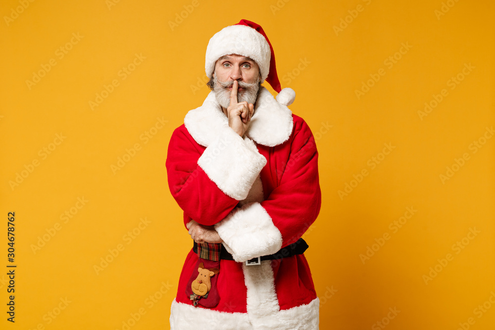 Elderly gray-haired mustache bearded Santa man in Christmas hat posing isolated on yellow background. New Year 2020 celebration concept. Mock up copy space. Say hush with finger on lips shhh gesture. Elderly gray-haired mustache bearded Santa man in Christmas hat posing isolated on yellow background. New Year 2020 celebration concept. Mock up copy space. Say hush with finger on lips shhh gesture.