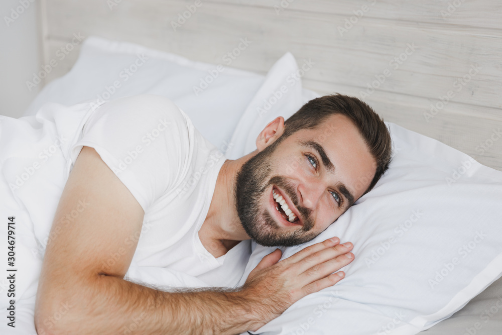 Calm young handsome bearded man lying in bed with white sheet pillow blanket in bedroom at home. Smiling beauty male spending time in room. Rest relax good mood lifestyle concept. Mock up copy space. Calm young handsome bearded man lying in bed with white sheet pillow blanket in bedroom at home. Smiling beauty male spending time in room. Rest relax good mood lifestyle concept. Mock up copy space.