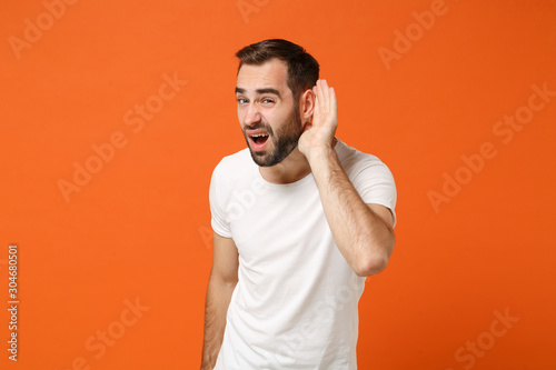 Concerned young man in casual white t-shirt posing isolated on orange background, studio portrait. People sincere emotions lifestyle concept. Mock up copy space. Try to hear you with hand near ear.