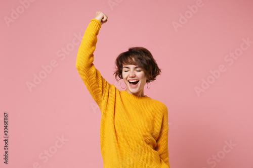 Joyful young brunette woman girl in yellow sweater posing isolated on pastel pink background studio portrait. People lifestyle concept. Mock up copy space. Doing winner gesture, keeping eyes closed. photo