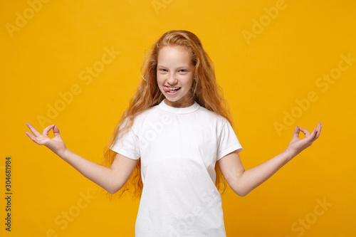 Funny little ginger kid girl 12-13 years old in white t-shirt isolated on yellow background in studio. Childhood lifestyle concept. Mock up copy space. Hold hands in yoga gesture, relaxing meditating.