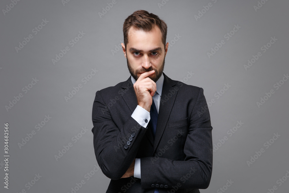 Serious young bearded business man in classic black suit shirt tie posing isolated on grey wall background. Achievement career wealth business concept. Mock up copy space. Put hand prop up on chin. Serious young bearded business man in classic black suit shirt tie posing isolated on grey wall background. Achievement career wealth business concept. Mock up copy space. Put hand prop up on chin.
