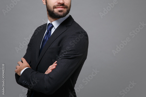 Cropped image of young bearded business man in classic black suit shirt tie posing isolated on grey background. Achievement career wealth business concept. Mock up copy space. Holding hands crossed.