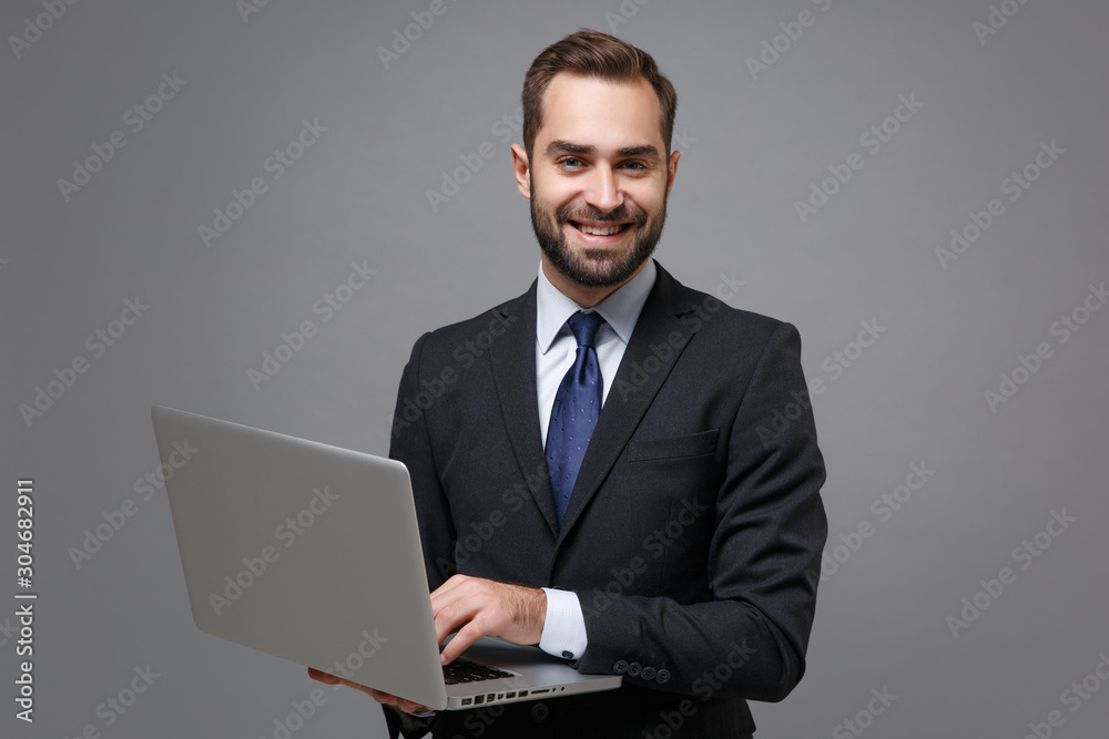 Smiling young business man in classic black suit shirt tie posing isolated on grey background. Achievement career wealth business concept. Mock up copy space. Holding, working on laptop pc computer. Smiling young business man in classic black suit shirt tie posing isolated on grey background. Achievement career wealth business concept. Mock up copy space. Holding, working on laptop pc computer.