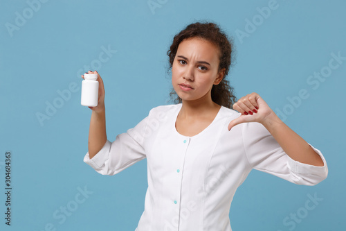 African american female doctor woman in medical gown showing thumb down holding tablets, aspirin pills in bottle isolated on blue background. Healthcare personnel medicine concept. Mock up copy space. © ViDi Studio