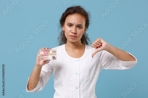 Displeased african american doctor woman isolated on blue background. Female doctor in medical gown showing thumb down hold daily pill box. Healthcare personnel medicine concept. Mock up copy space.