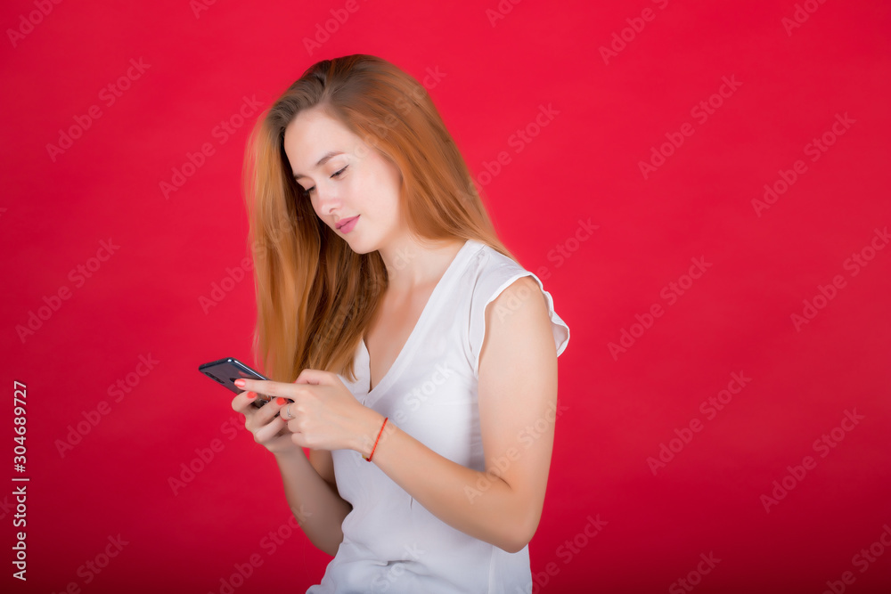Young, beautiful red-haired woman isolated on a red background, looks at the phone screen. Young, beautiful red-haired woman isolated on a red background, looks at the phone screen.