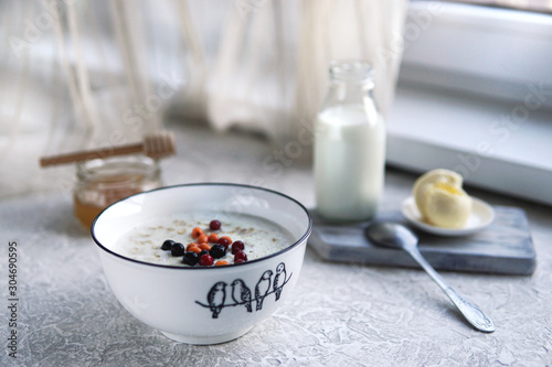 Bowl of oatmeal porridge with berries, honey, bottle of milk, butter on grey table near window, hot and healthy food for Breakfast, selective focus.