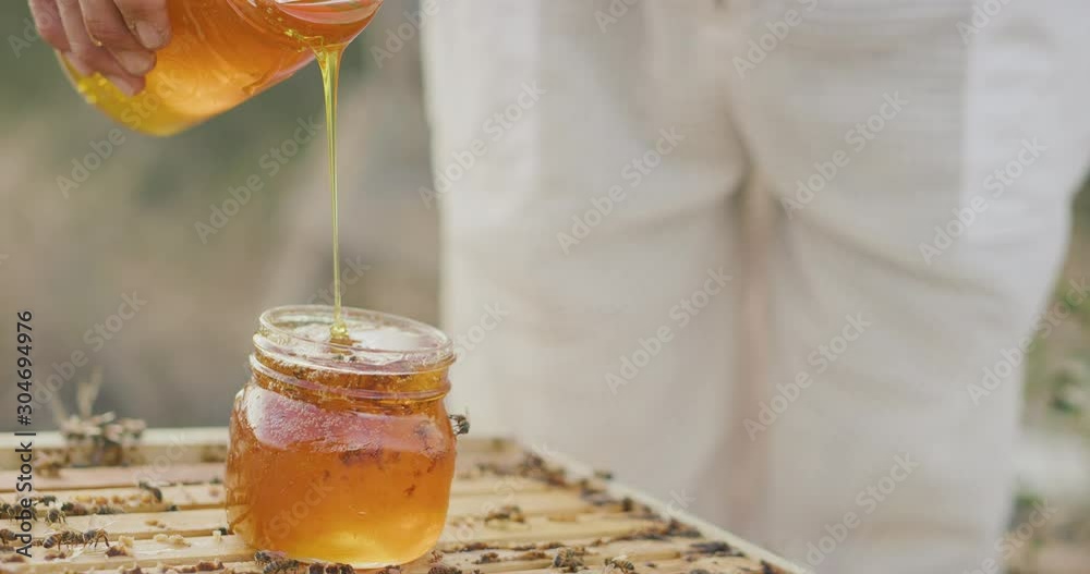 Close up shot of a beekeeper pouring golden honey into a glass jar on top of a beehive with buzzing bees, beekeeper harvesting honey