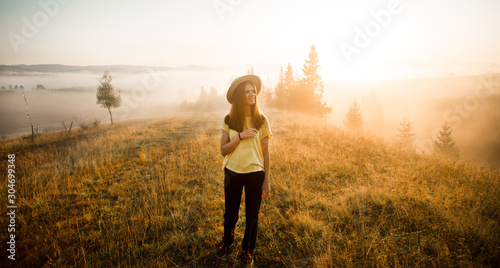 Carefree happy woman in yellow shirt and straw hat enjoying nature on grass meadow on top of mountain with sunrise. Beauty girl outdoor with sunbeams. Freedom concept