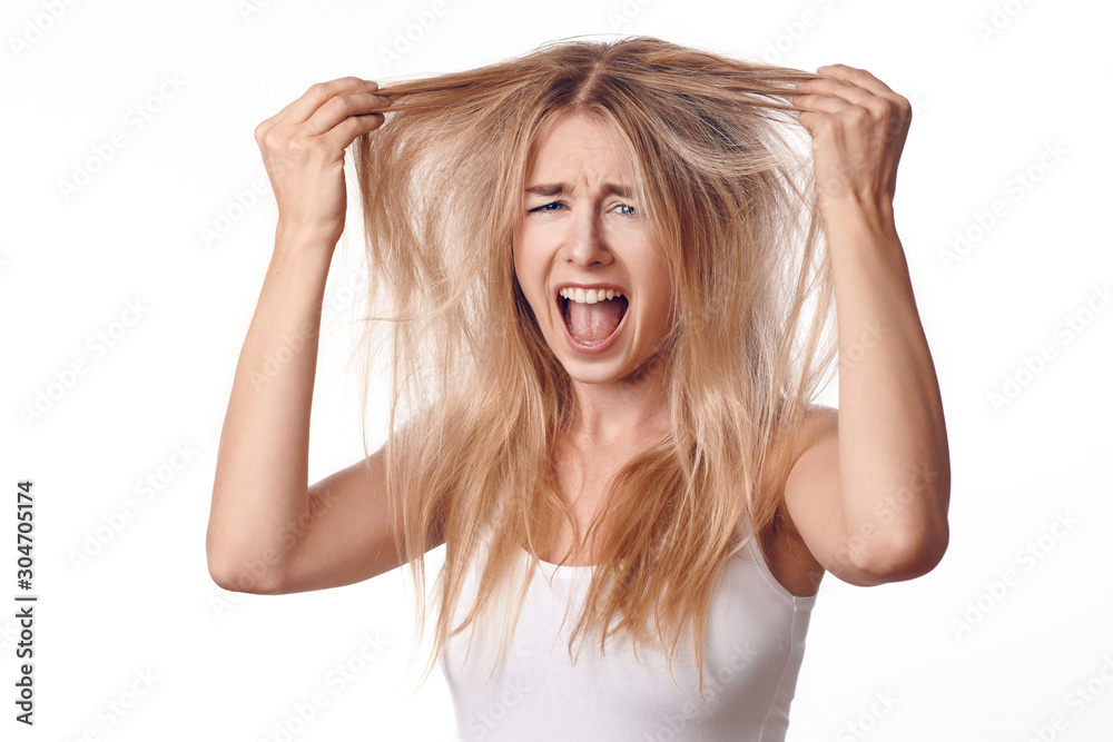 Young blond woman unhappy with her haircut, touching her hair with both hands and looking at camera with screaming grimace. Front portrait on white background Young blond woman unhappy with her haircut, touching her hair with both hands and looking at camera with screaming grimace. Front portrait on white background