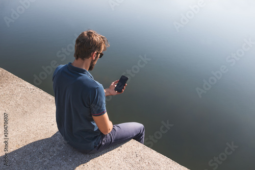 Young caucasian man using his cell phone while sitting on the lake shore