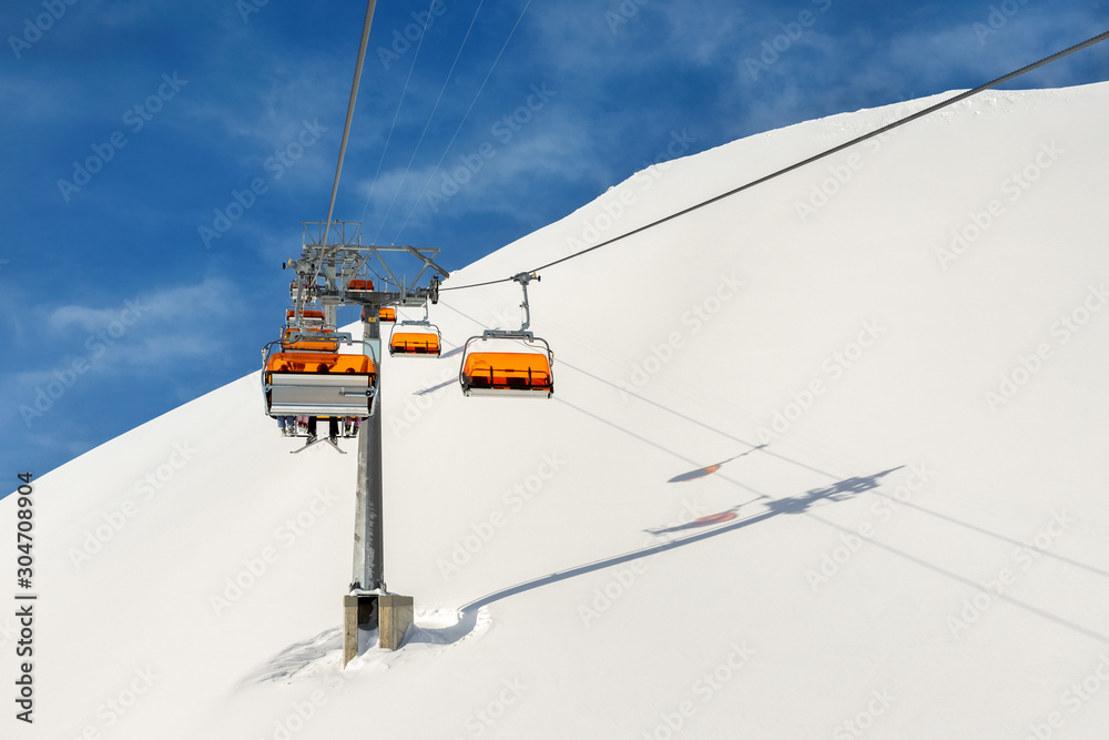 Ski lift ropeway on hilghland alpine mountain winter resort on bright sunny day. Ski chairlift cable way with people enjoy skiing and snowboarding.Banner panoramic wide view of downhill slopes Ski lift ropeway on hilghland alpine mountain winter resort on bright sunny day. Ski chairlift cable way with people enjoy skiing and snowboarding.Banner panoramic wide view of downhill slopes