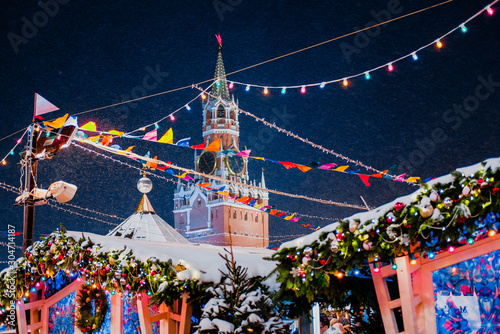 Spasskaya tower of Kremlin among New Year festive decorations on Red Square, main landmark in Moscow. Christmas fair in Russia at evening while snow falling.