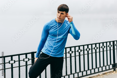 Young jogger muscular man stretching outdoor in the morning on the fog nature background. Fitness male exercising in the park and listenting the music on earphones. People and sport concept photo
