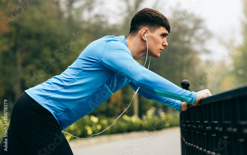 Side view image of young sportsman stretching outdoor in the morning on the fog nature background. Fitness male exercising in the park and listenting the music on earphones. People and sport concept photo