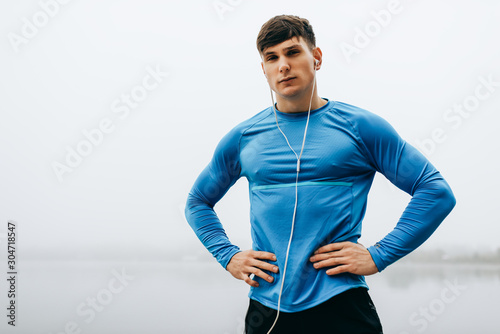 Image of young athlete man posing outdoor in the morning on the fog nature background during running. Fitness male exercising in the park and listenting the music on earphones. People, sport concept photo