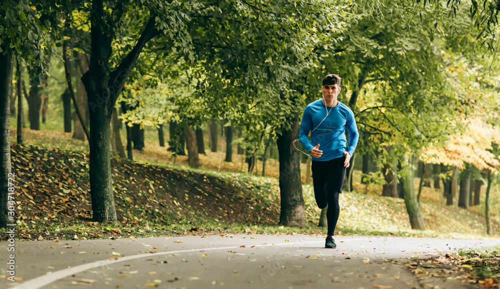 Fototapeta premium Horizontal full body image of young jogger man jogging outdoor in the forest background. Fitness male exercising in the park, wearing blue and black sportswear. People and sport concept