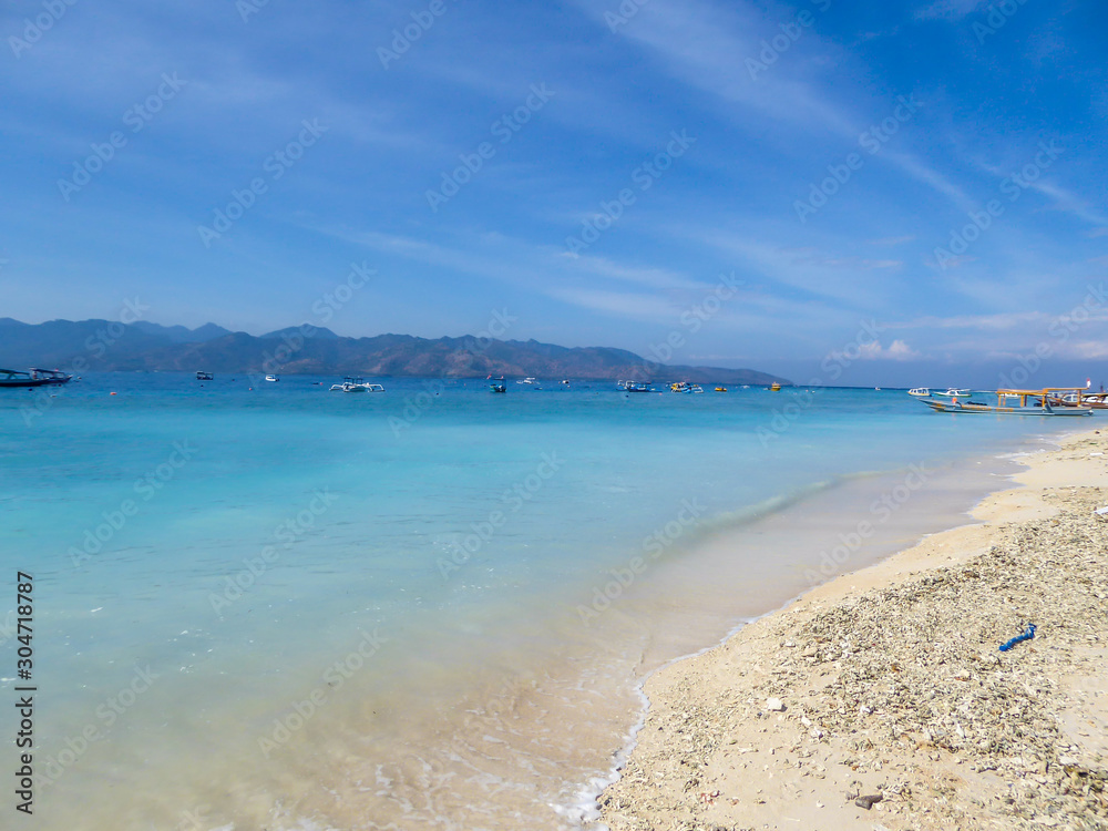 Lots of boats anchored next to the shore of Gili Air, Lombok Indonesia. Beautiful and clear water. In the back visible Mount Rinjani. Some trees on the shore, few clouds on the sky. Holidays paradise