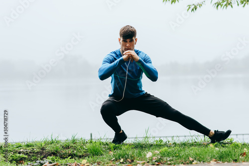Outdoors horizontal image of athletic man stretching outdoor in the morning on the fog nature background. Fitness male exercising in the park and listenting the music on earphones. People and sport photo