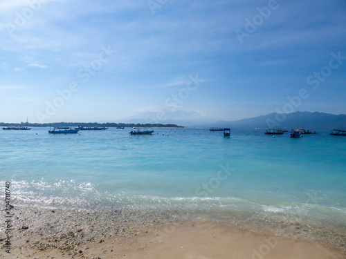 Lots of boats anchored next to the shore of Gili Air, Lombok Indonesia. Beautiful and clear water. In the back visible Mount Rinjani. Some trees on the shore, few clouds on the sky. Holidays paradise © Chris