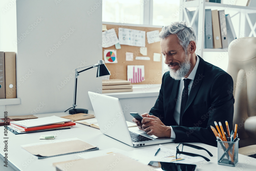 Thoughtful mature man in full suit smiling and using smart phone while working in modern office
