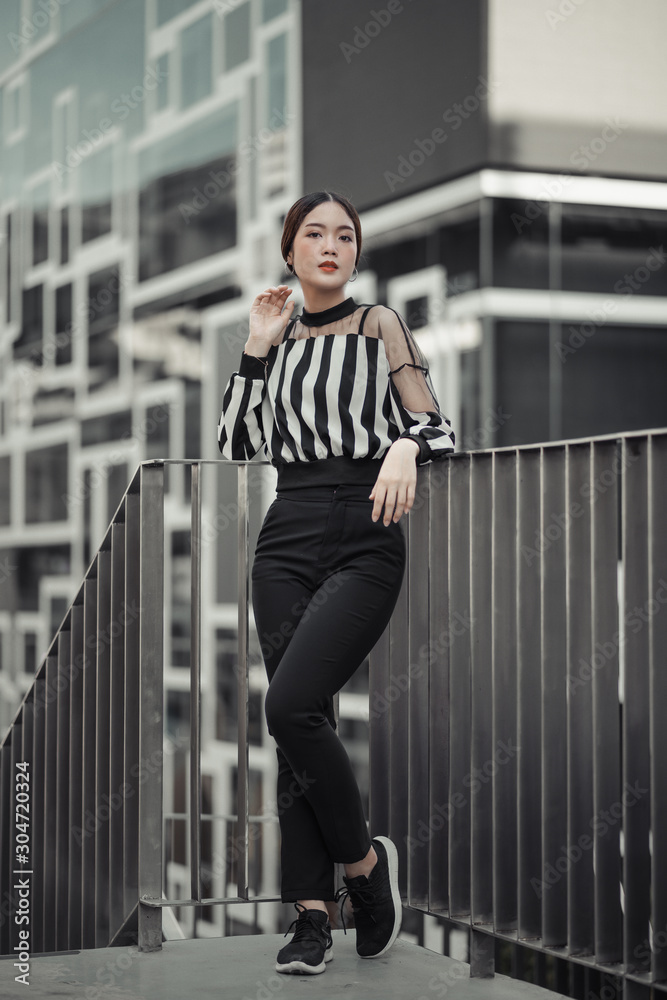 street portrait, strong woman in black, confident woman, lady in city, portrait of Asian female, standing and hand on armrest against the railing, young lady in black white stripe top