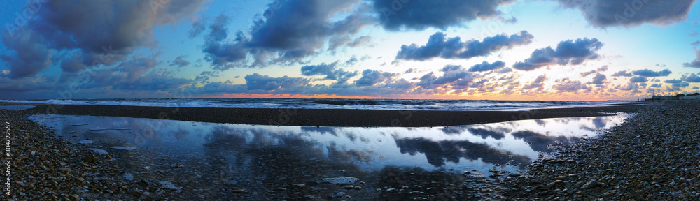 Obraz premium Awesome panoramic seascape at sunset at gravelly beach with sandbanks to form a natural pool where the clouds are reflected in the water and sky have marvelous orange and dark blue twilight