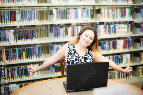 Beautiful white woman female student smiling and stretching body for relaxing while working with laptop computer at her desk, office lifestyle, business situation. Bookshelves in library