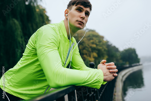 Horizontal close-up portrait of young athletic man resting in the morning after workout on the fog lake background. Fitness male exercising in the park and listenting the music on earphones. photo