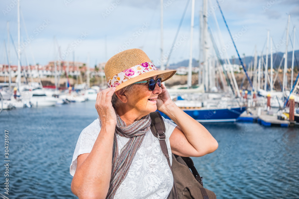 Portrait of a senior attractive  woman with gray hairs and hat sitting at the port. A small port with yachts and sailboats. Cloudy and windy day