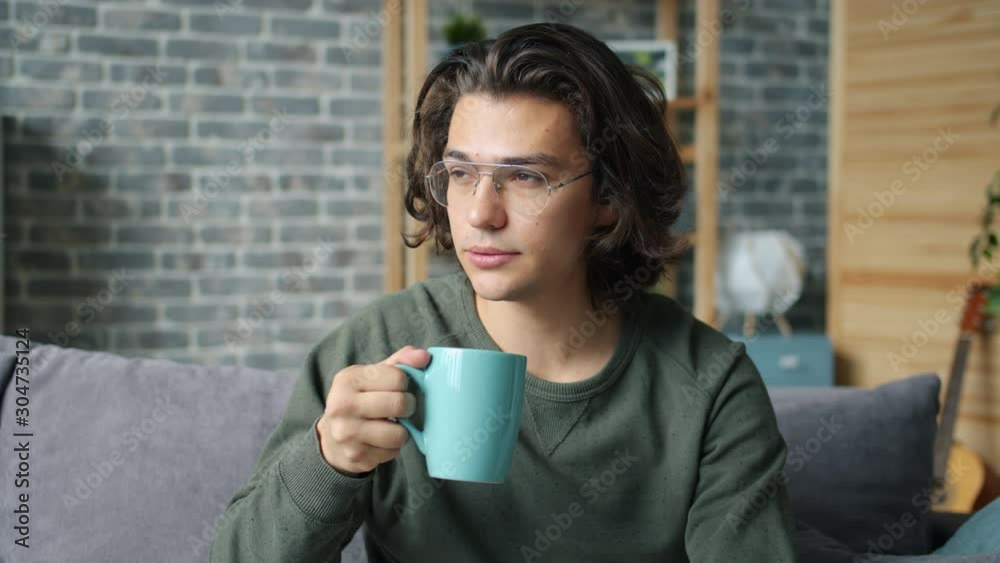 Slow motion of attractive young man in glasses drinking from mug at home relaxing sitting on couch alone. Drinks, modern lifestyle and millennials concept. Slow motion of attractive young man in glasses drinking from mug at home relaxing sitting on couch alone. Drinks, modern lifestyle and millennials concept.