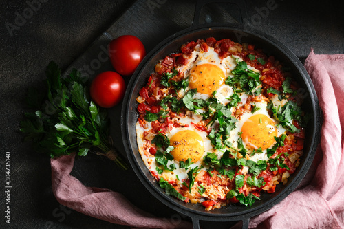 Traditional  shakshuka with eggs, tomato, and parsley in a iron pan on a dark background. Top view.