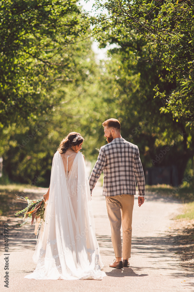 Happy bride and groom holding hands and walking in garden wedding day. Back view of charming stylish newlyweds holding hands while walking in park forest. Path to future life. happy marriage moments Happy bride and groom holding hands and walking in garden wedding day. Back view of charming stylish newlyweds holding hands while walking in park forest. Path to future life. happy marriage moments