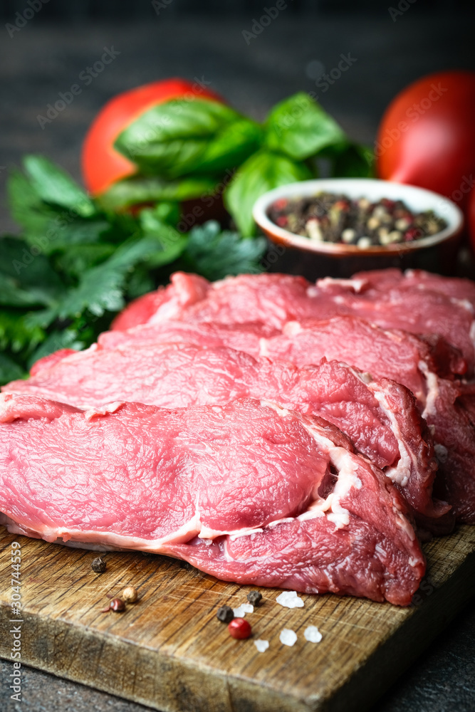 Fresh raw beef, cut into steaks on a wooden board with vegetables, herbs and spices on a wooden board on a dark background. Fresh raw beef, cut into steaks on a wooden board with vegetables, herbs and spices on a wooden board on a dark background.
