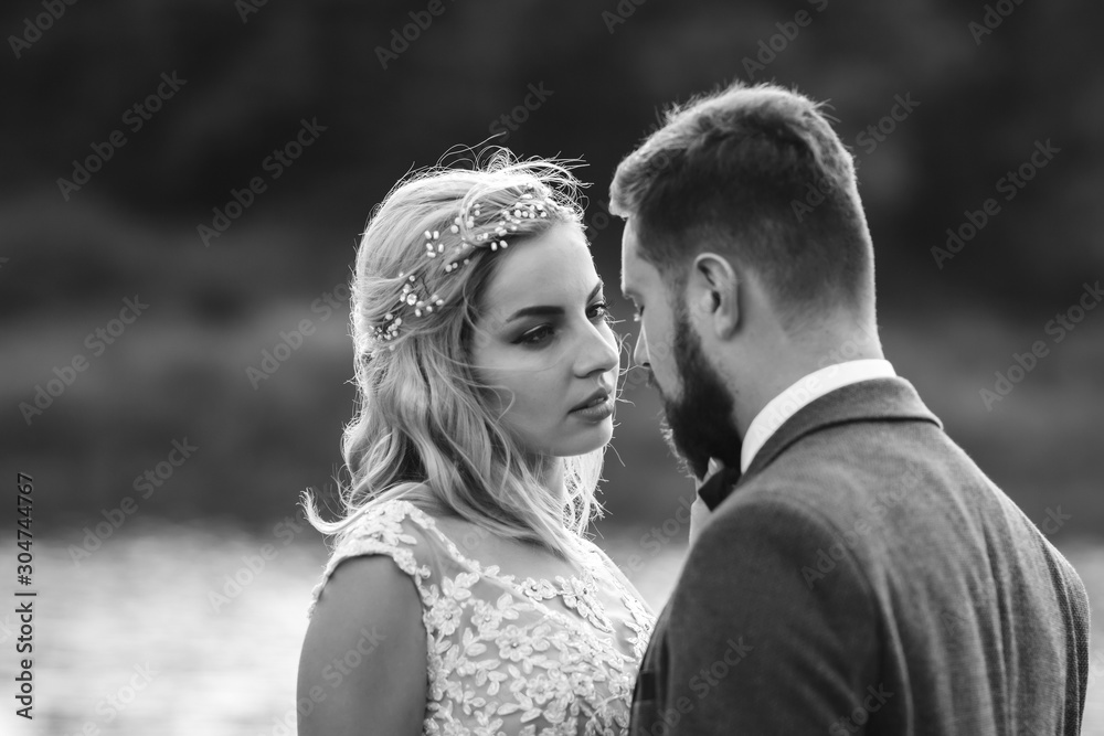 Black and white foto stylish couple of newlyweds posing on a bridge on wedding day. Handsome bearded groom admires and kisses pretty bride. Together. The concept of youth, love, fashion and lifestyle.